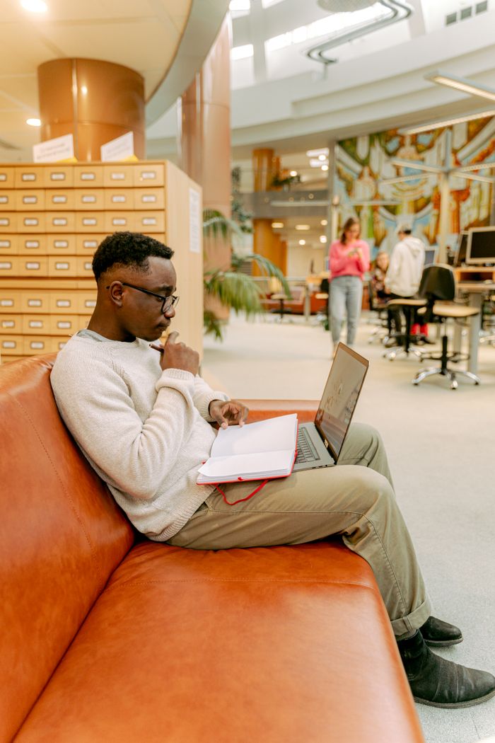 Services African American man using laptop and notebook in a library setting, focused on research.