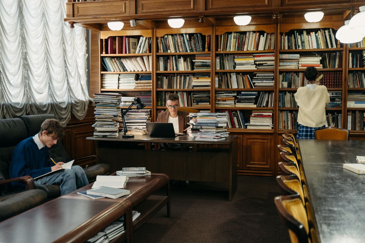 Home Students studying in a spacious library, surrounded by wooden shelves filled with books.